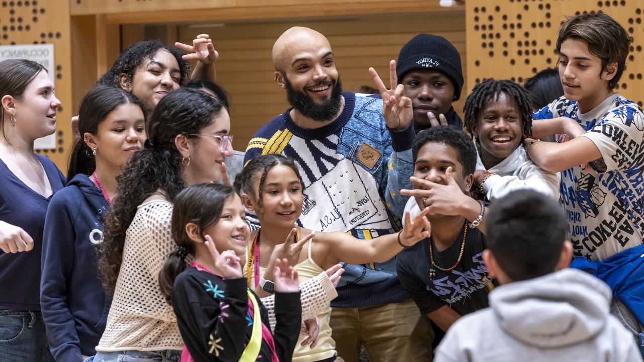A joyful moment among young students gathered around 马库斯·诺里斯 in an orchestral rehearsal studio at Juilliard. Norris is smiling broadly and wearing a stylish, patterned sweater. 他站在中间, surrounded by the enthusiastic students posing playfully—some making peace signs, while others smile or gesture animatedly towards the camera.